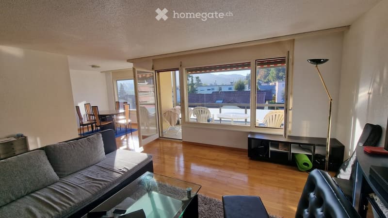 Living room with a grey sofa, coffee table, dining area with chairs and table, large windows with a view, and a balcony area with outdoor furniture.