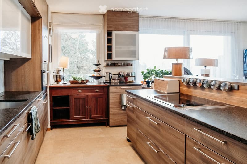 Modern kitchen with wooden cabinets, stainless steel appliances, granite countertops, and a large window with sheer curtains. Includes a sink, oven, microwave, refrigerator, blender, coffee maker, toaster, and decorative items on the counter.