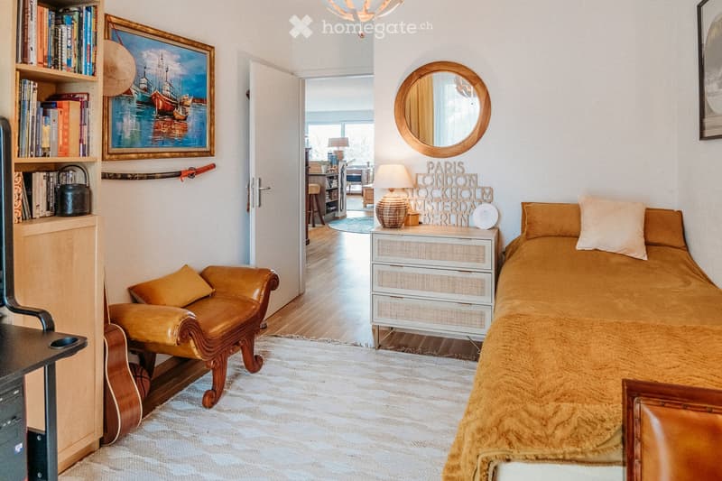 Cozy bedroom with a bed, leather armchair, nightstand with lamp, bookshelf, and a round mirror on the wall. The room features warm tones and a mix of modern and vintage decor.