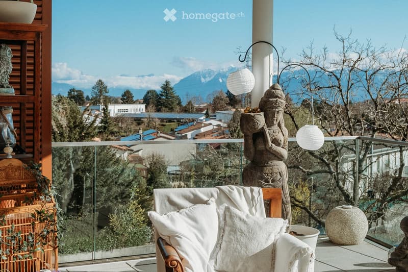 Balcony with a view of mountains, featuring a wooden chair with white cushions, a decorative statue, a hanging lantern, and a ceramic vase.