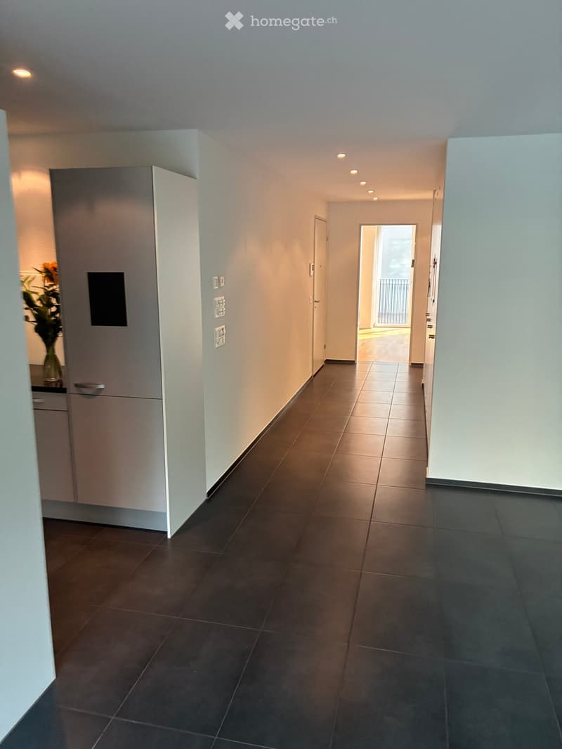Modern hallway with tiled flooring, white walls, recessed lighting, and a small cabinet with a vase of flowers on the left side. A doorway leads to a balcony area with a railing visible.