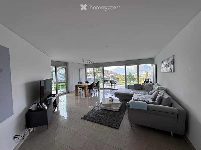 Modern living room with large windows offering a scenic view, featuring a grey sectional sofa, coffee table, side table, dining table, and chairs.