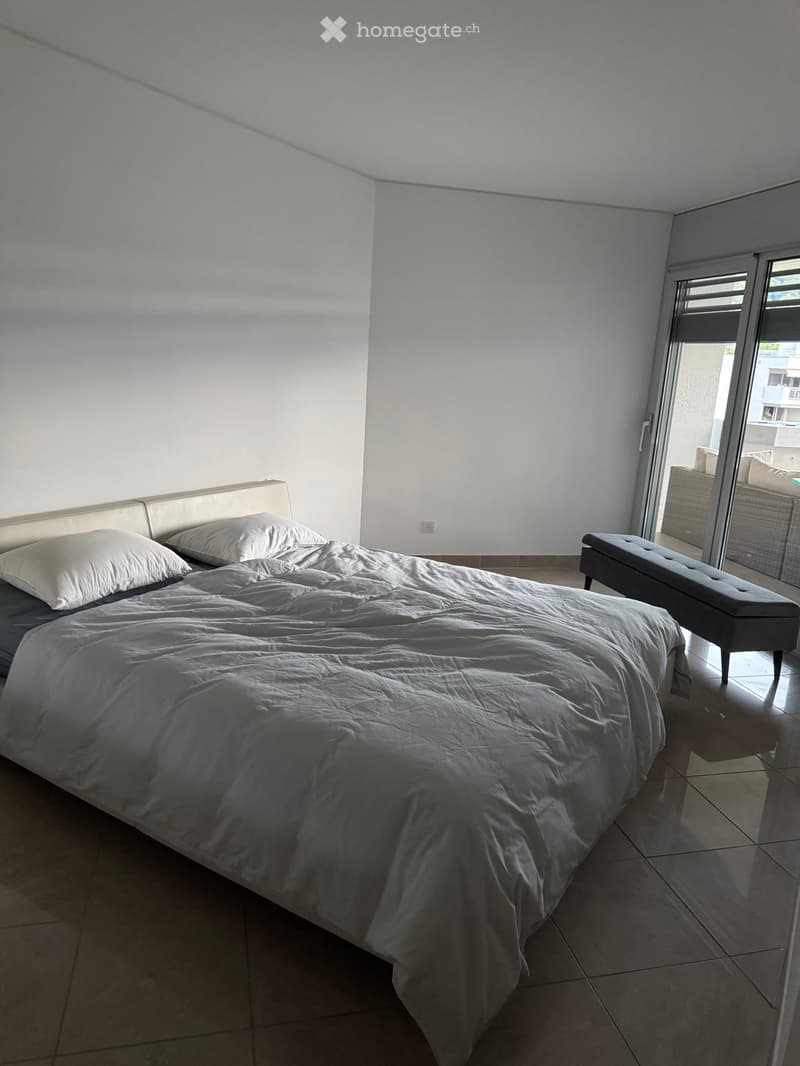 Modern bedroom with a large bed, pillows, and a bench near a sliding glass door leading to a balcony.