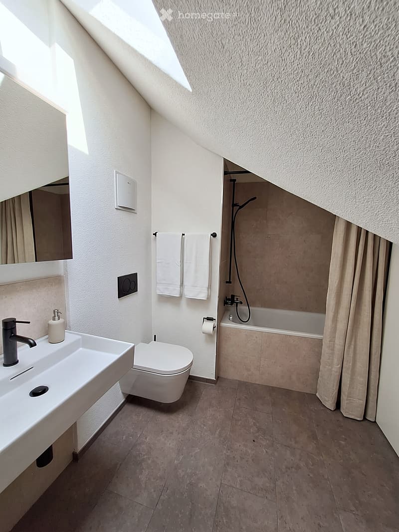 Modern bathroom with white fixtures, a sink, toilet, bathtub, and towels hanging on a rack. The room features a skylight and a sleek, contemporary design.