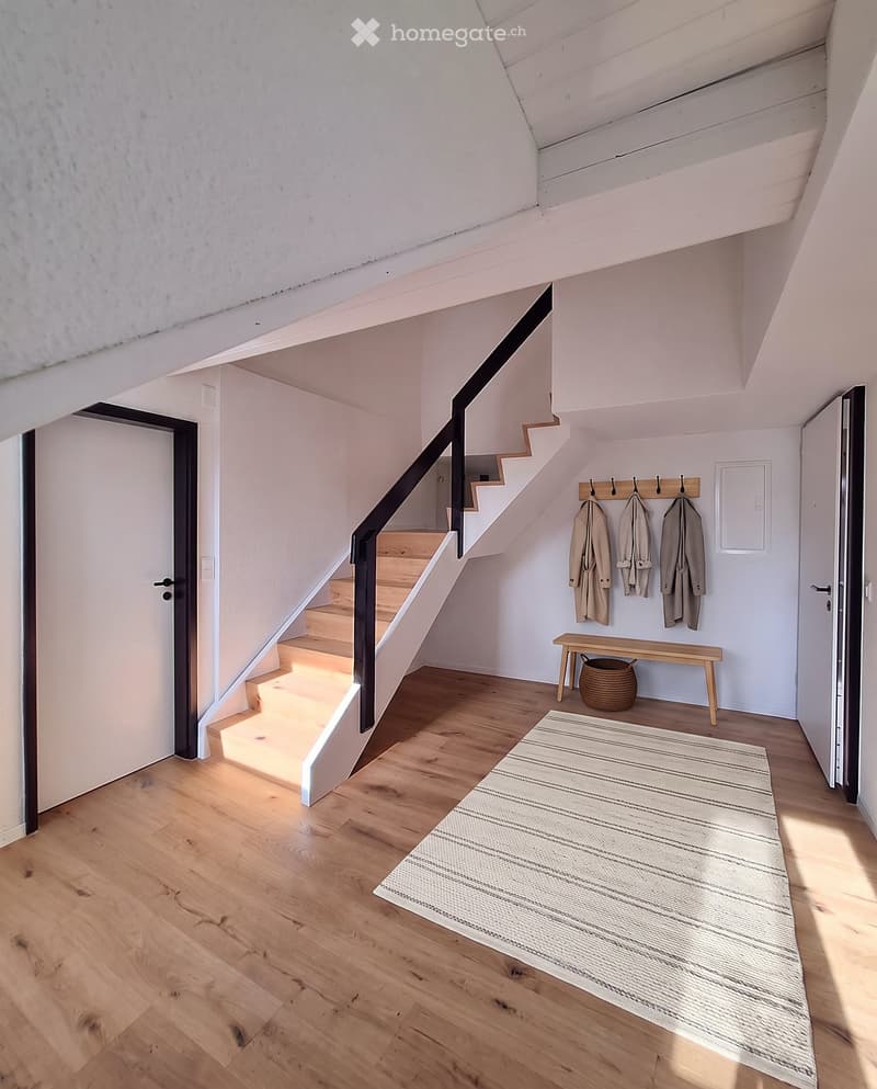 Bright hallway with wooden flooring, a staircase leading upwards, a small bench, a woven basket, and coats hanging on a rack.