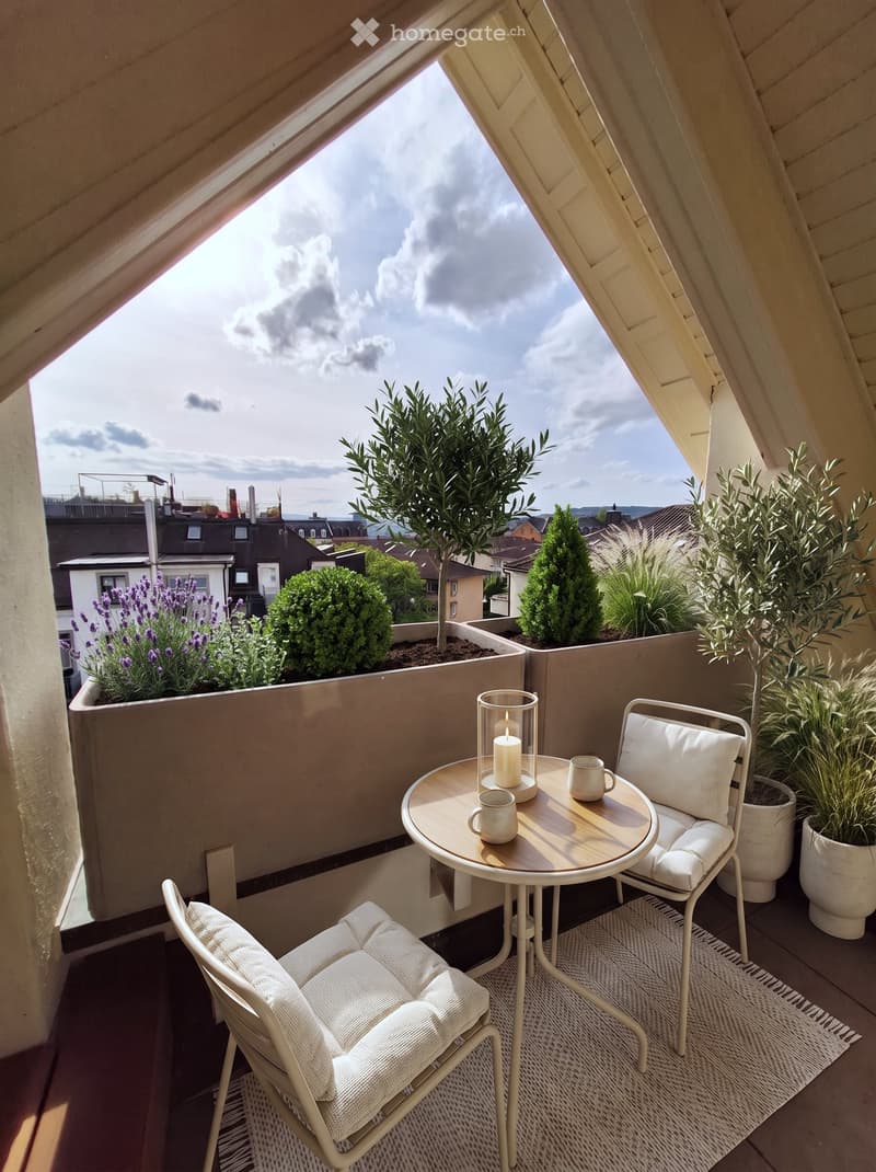 A small, cozy balcony with a round wooden table, two white chairs, a candle in a glass holder, and two cups. Potted plants and flowers are arranged along the edge, with a view of the cityscape.