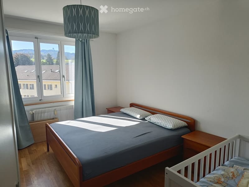 Cozy bedroom with a wooden bed, nightstands, a small cradle, and a radiator beneath a large window with a scenic view.