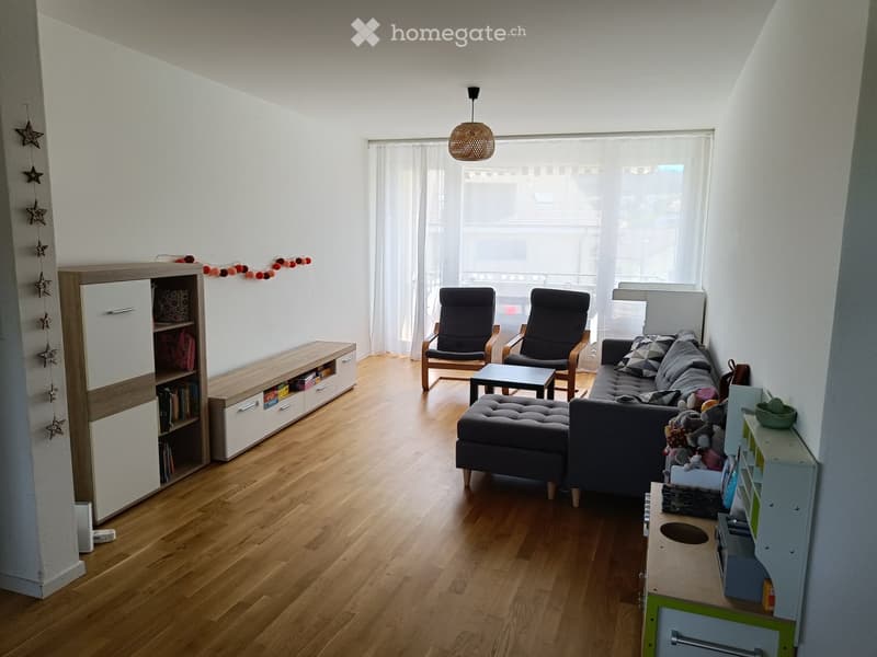 Bright living room with wooden flooring, large window with white curtains, grey sofa with ottoman, two armchairs, coffee table, bookcase, and children's play area.