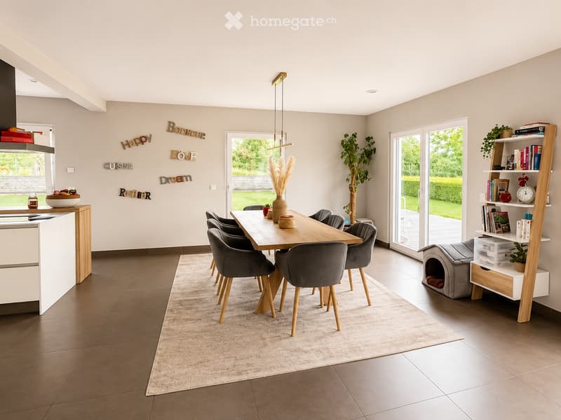Modern dining area with a wooden dining table, six grey chairs, a bookshelf, a cat bed, and decorative items including a vase and potted plants. The space features large windows with a view of a green garden.