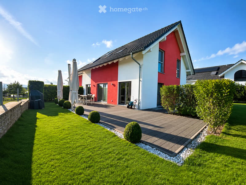 A modern two-story house with red and white exterior walls, featuring a well-maintained lawn, a wooden deck, patio furniture, and a barbecue area.