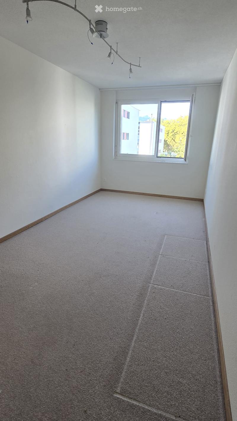 Empty room with light-colored walls, carpeted floor, and a single window with white frame. No furniture or equipment visible.