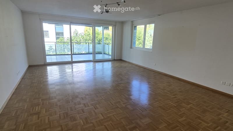 Empty room with wooden flooring, white walls, large windows, and sliding glass doors leading to a balcony.