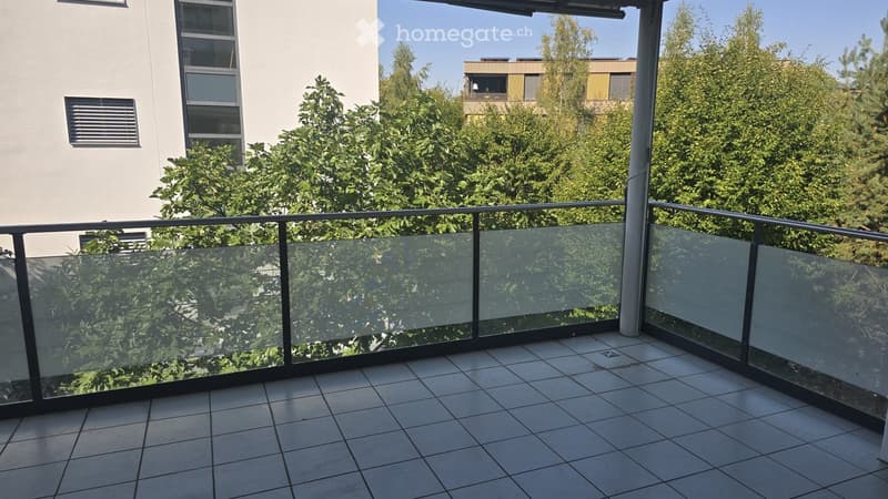 Balcony with tiled floor, glass railing, and view of greenery and neighboring buildings.