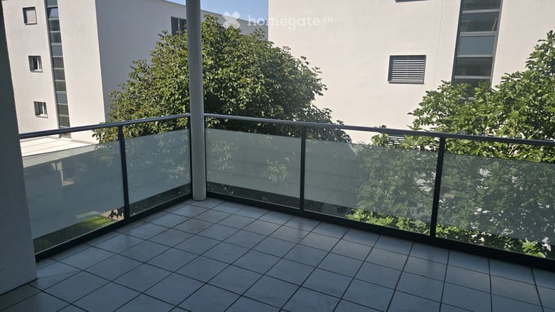 Balcony with tiled floor and glass railing, overlooking a modern building and greenery.