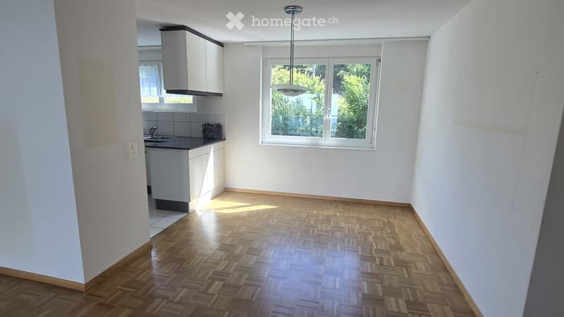 Empty room with wooden herringbone flooring, white walls, and a large window with a view of greenery. The adjacent kitchen area features white cabinets, a sink, and a countertop.