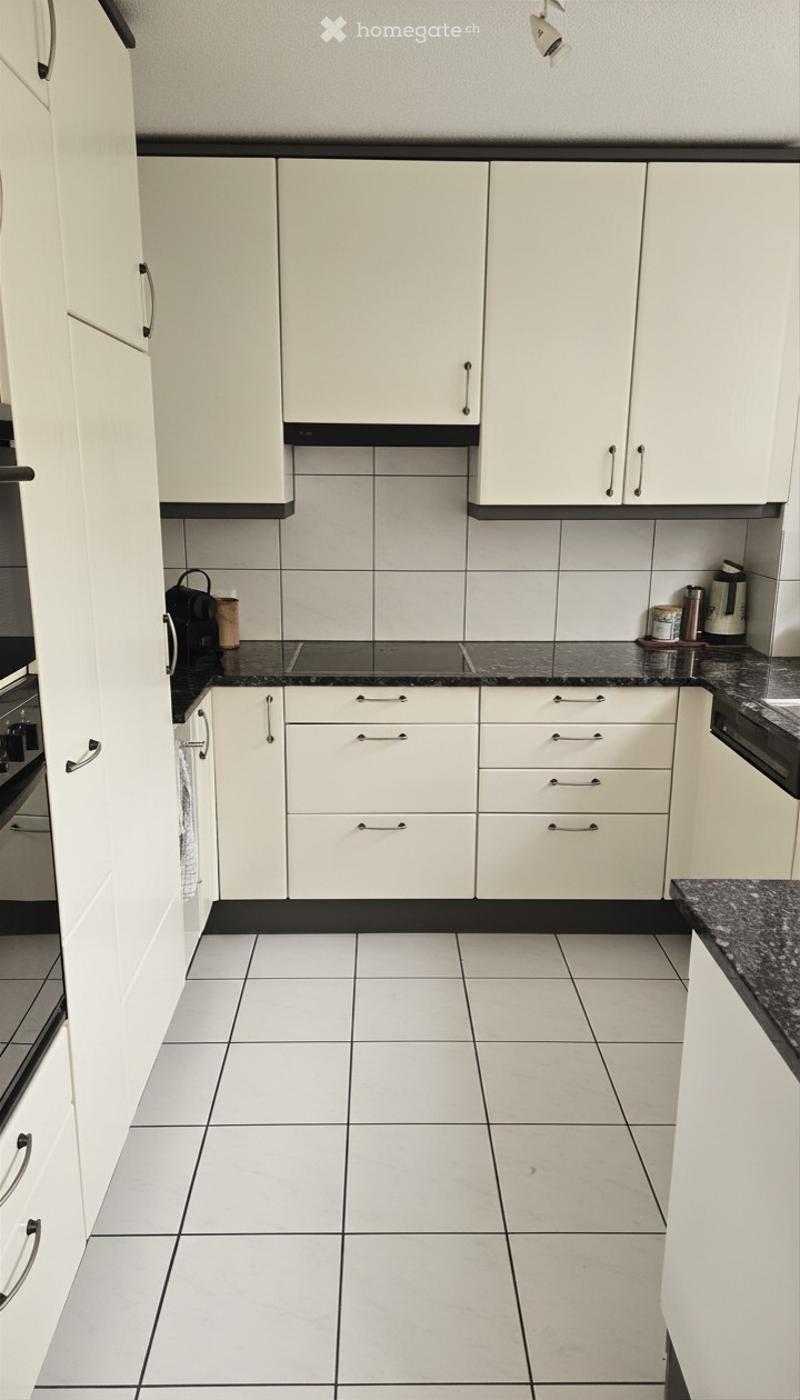 Modern kitchen with white cabinets, black countertops, and tiled flooring. Includes an oven, coffee maker, and various small appliances on the counter.