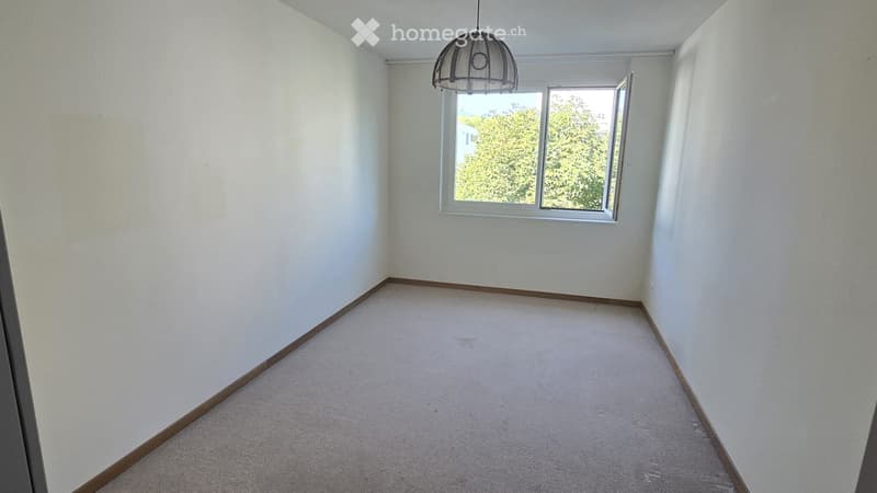 Empty room with white walls, carpeted floor, and a single window with a hanging light fixture above it.