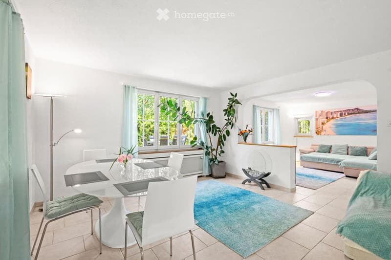 Bright, modern living and dining area with white walls, large windows, a sofa, dining table, chairs, a fireplace, and decorative plants.