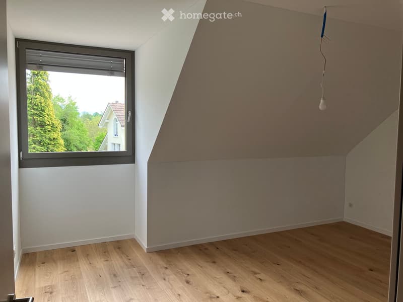 Empty attic room with wooden flooring, white walls, and a large window offering a view of greenery and a neighboring house.