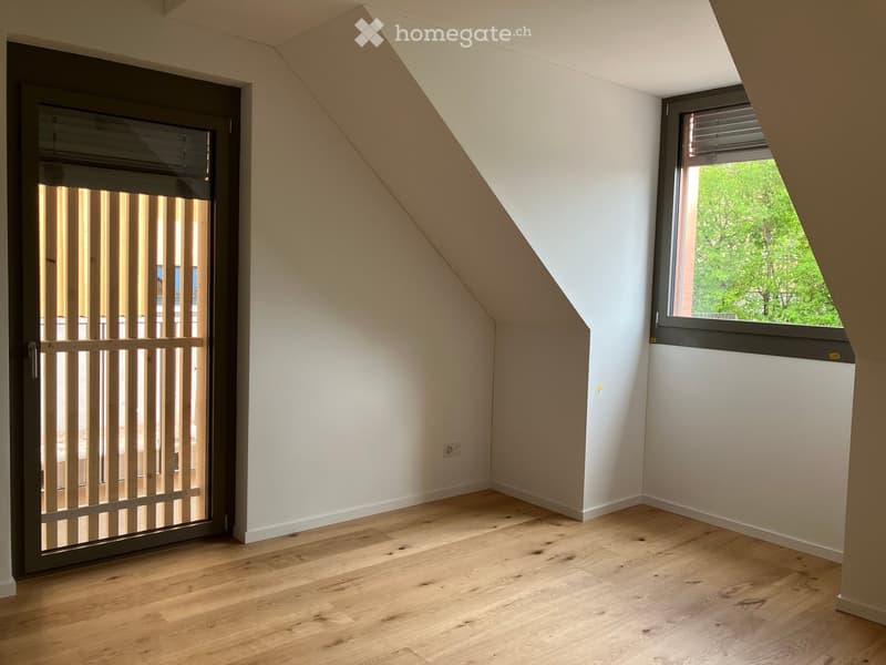 Empty attic room with wooden flooring, white walls, and a large window with blinds. The room features a door with a security gate and a slanted ceiling typical of attic spaces.