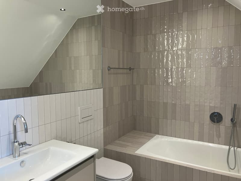 Modern bathroom with a white sink, bathtub, toilet, and showerhead. Features grey tiled walls and a sleek chrome faucet.