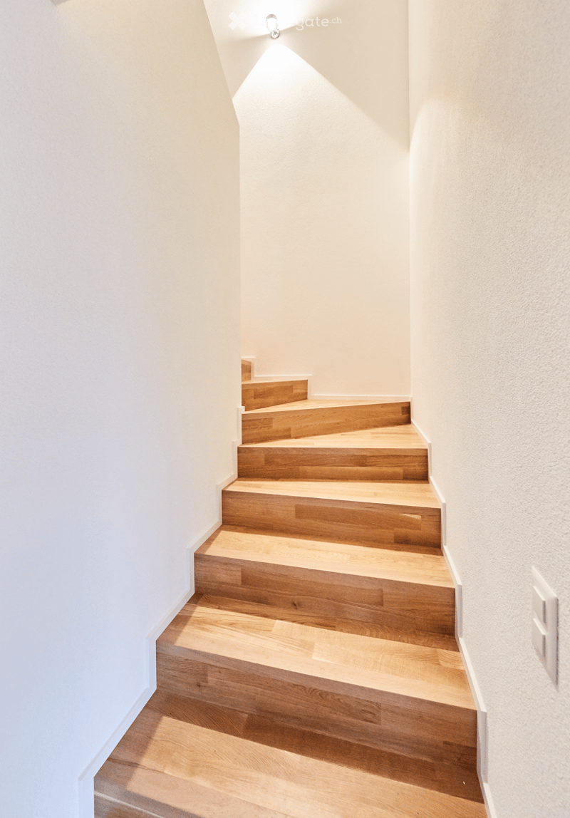 Modern staircase with wooden steps and white walls, illuminated by a wall-mounted light fixture.