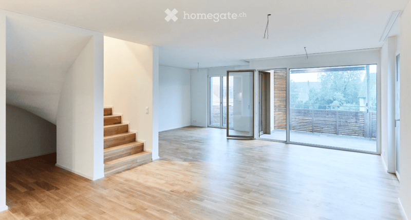 Bright, open room with wooden flooring, white walls, and large glass sliding doors leading to a balcony. A small wooden staircase is visible on the left side.