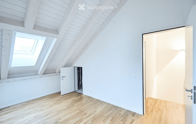 Empty attic room with wooden flooring, white walls, and a skylight window. The room features exposed wooden beams on the ceiling and a door leading to another space.