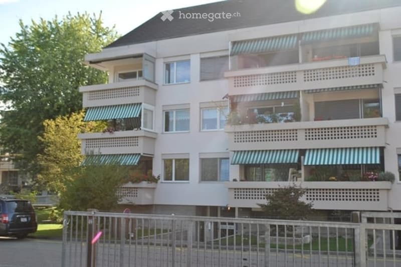 A multi-story residential building with balconies, large windows, and striped awnings. The building is surrounded by greenery and has a gated entrance.