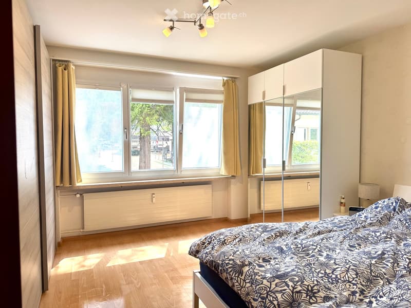 Bright bedroom with wooden flooring, large windows, a bed with patterned bedding, a wardrobe with mirrored doors, and a lamp on a side table.