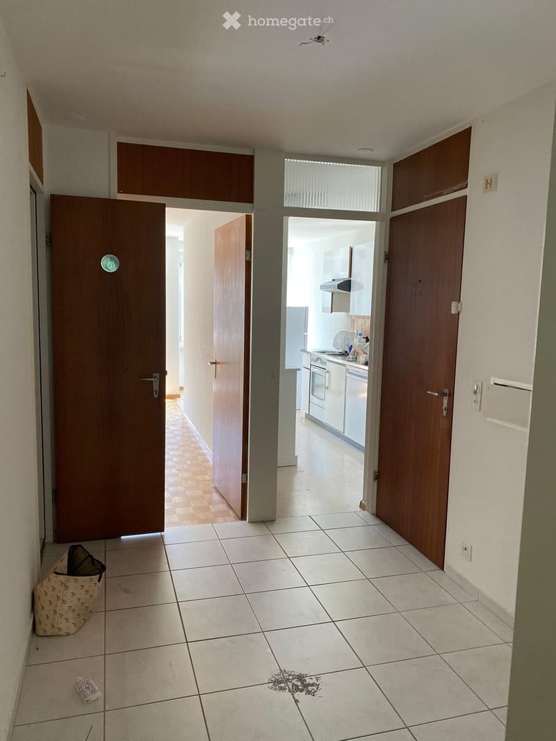 Empty hallway with tiled floor, wooden doors, and a view into a kitchen area with white cabinets and appliances.