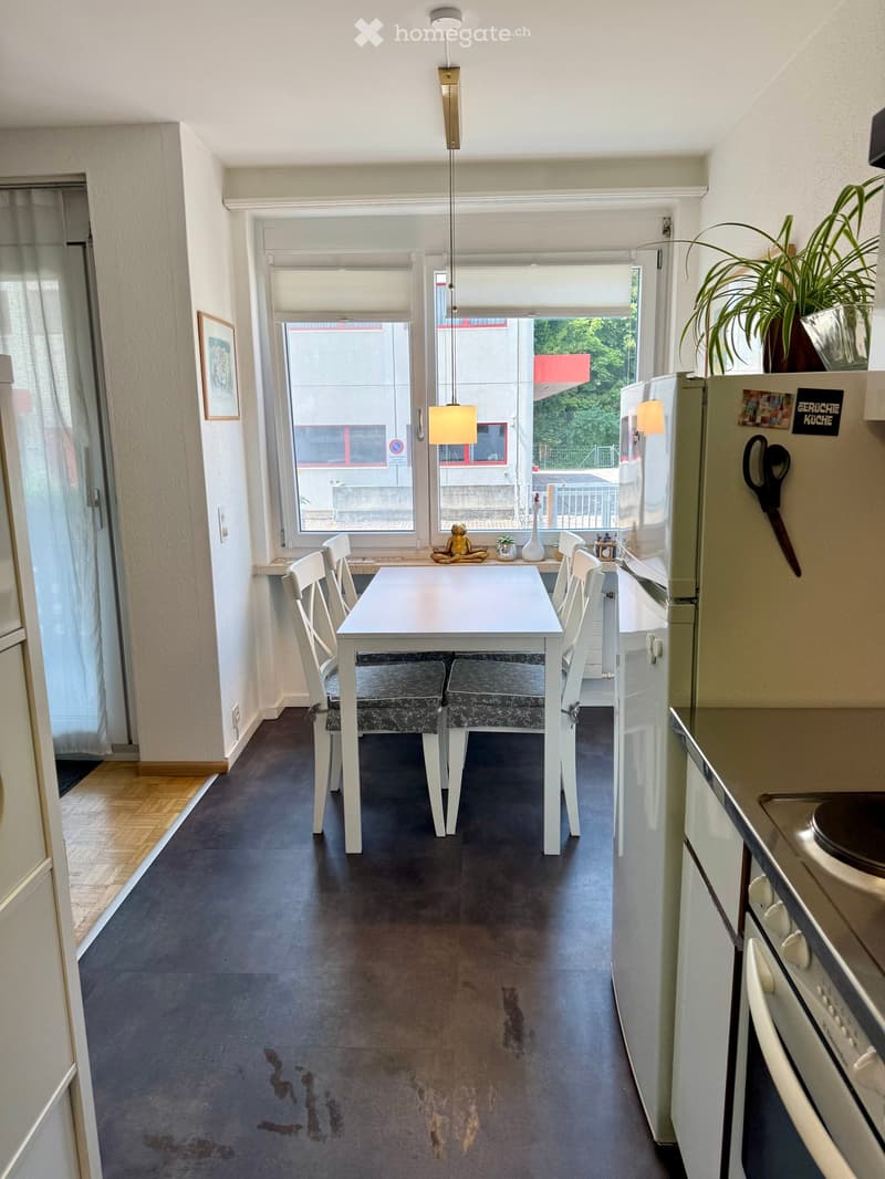 A modern kitchen and dining area with a white dining table, four chairs, two stools, a refrigerator, oven, and dishwasher. The space features large windows with blinds and a view of the outside, along with a potted plant and a decorative vase on the windo