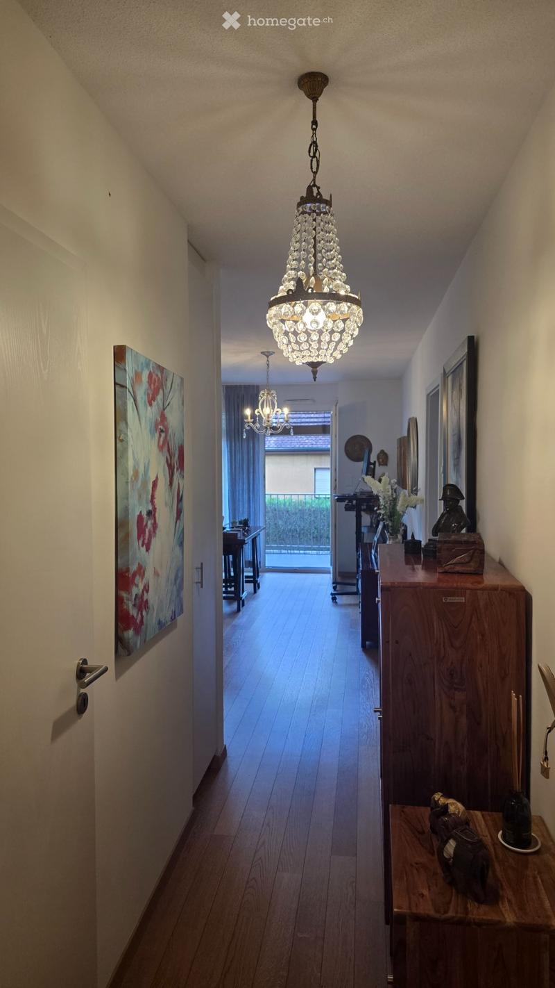 A well-lit hallway with wooden flooring, a chandelier, and a view into a room with a table and chairs. The hallway features a painting on the left wall and a small table with decorative items on the right.