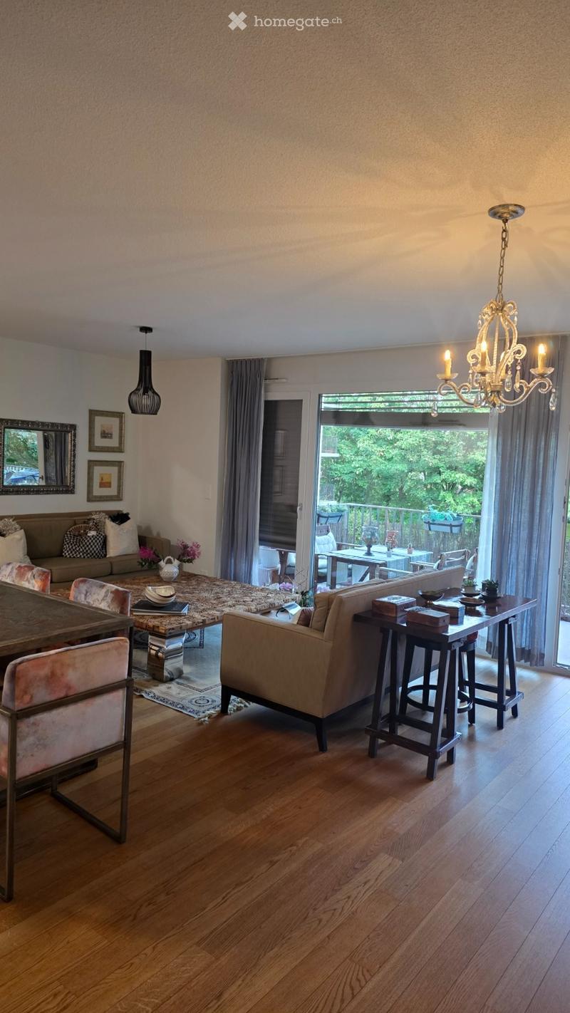 A spacious living room with wooden flooring, a large central dining table, a sofa, chairs, and a balcony view. The room features a chandelier, a side table with decorative items, and a mirror on the wall.