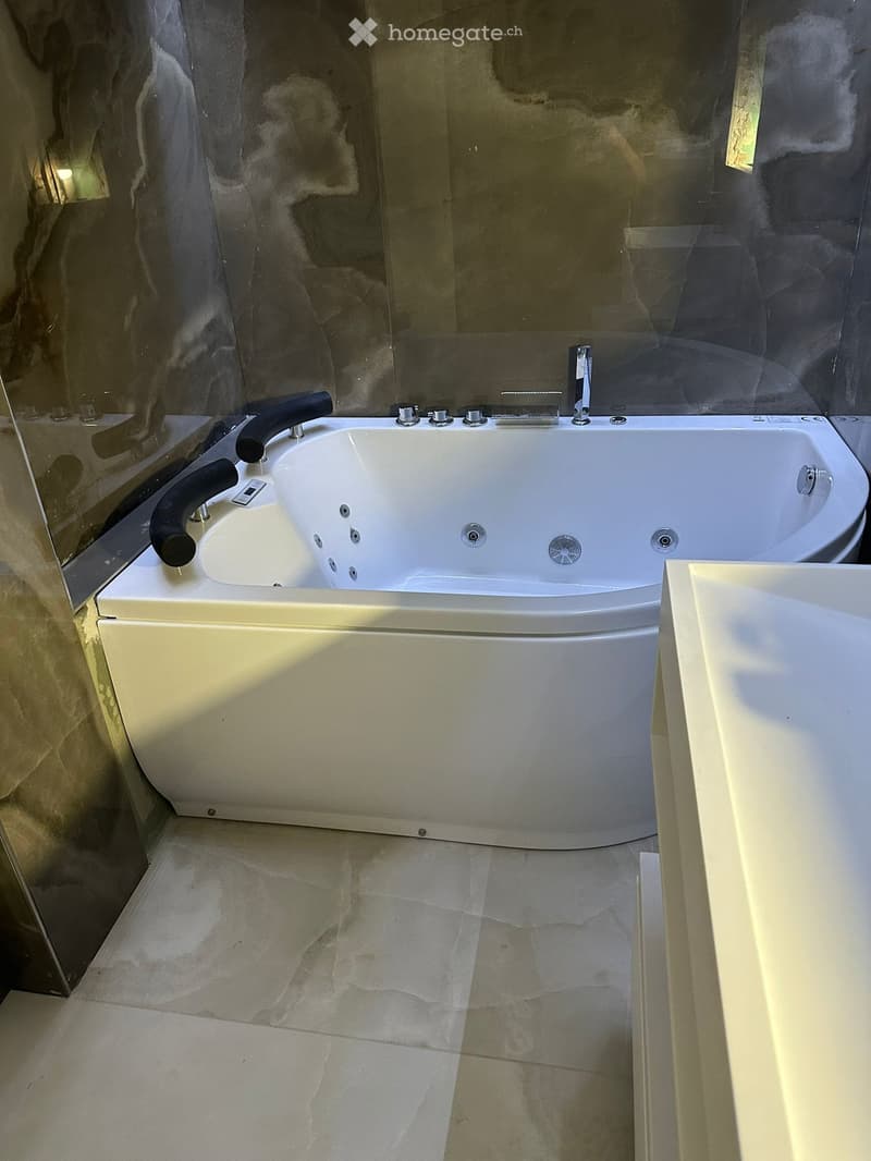 Modern bathroom with a large, white bathtub featuring multiple jets and a sleek faucet. The floor is tiled with light-colored stone, and the walls are partially covered with dark, textured stone panels.