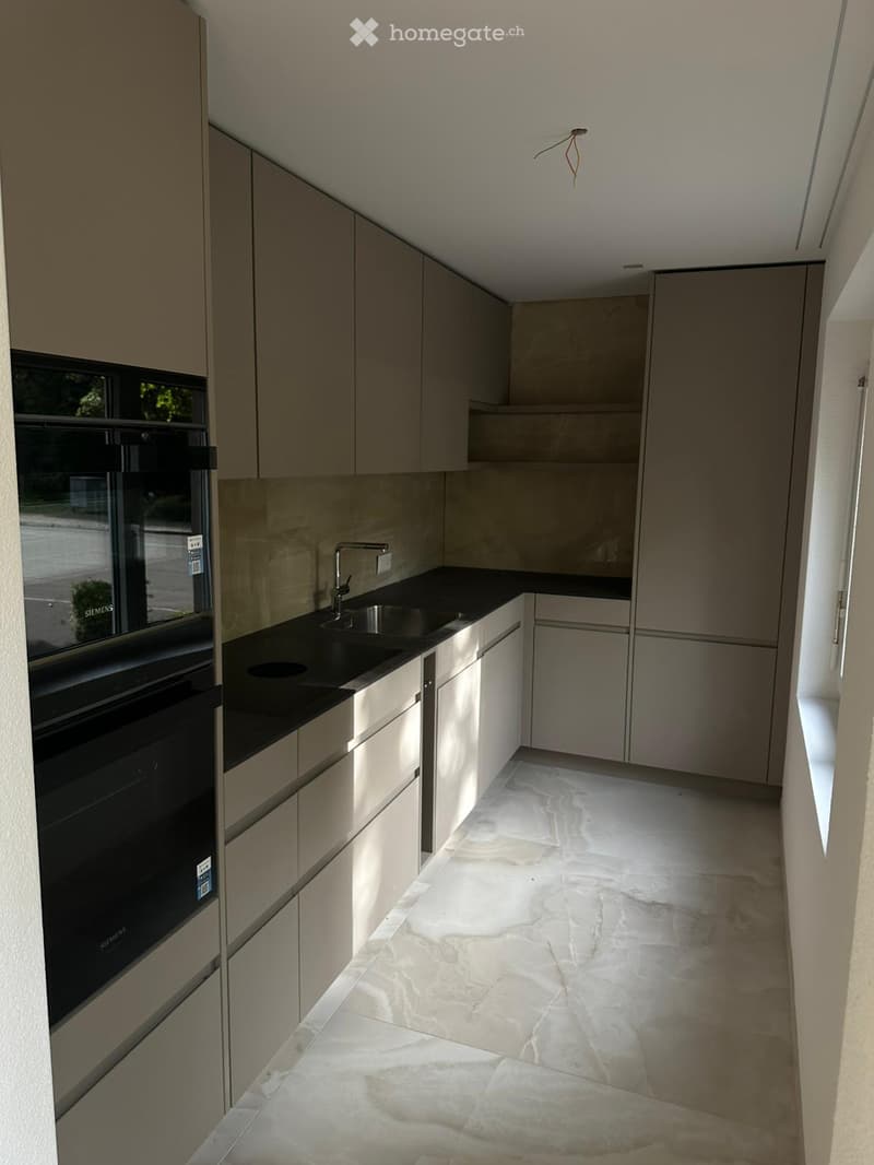 Modern kitchen with light grey cabinets, marble flooring, and a black countertop. Features a built-in oven and a sink with a modern faucet.