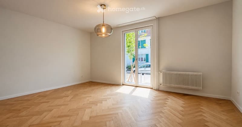 Empty room with wooden herringbone flooring, white walls, large glass sliding door leading to a balcony, and a modern pendant light fixture.