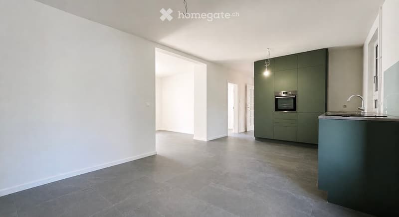 Empty room with grey tiled floor, white walls, and a small kitchen area with green cabinets, sink, and oven.