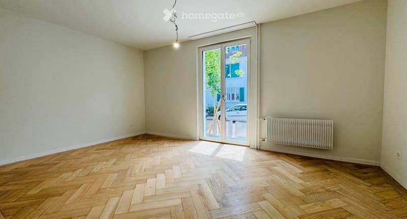 Empty room with wooden herringbone flooring, white walls, large glass sliding door leading outside, radiator on one wall, and a single hanging light fixture.