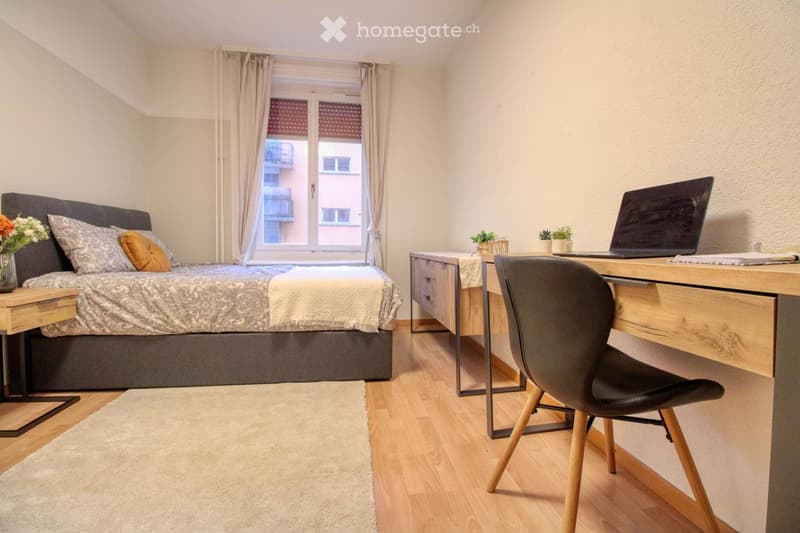 Bedroom: double bed with grey bedspread, wooden floor, white walls, wooden desk with laptop, black chair, wooden nightstand, and a window with curtains.