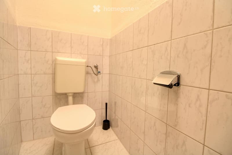 A small bathroom with white tiled walls, a toilet, and a toilet brush.