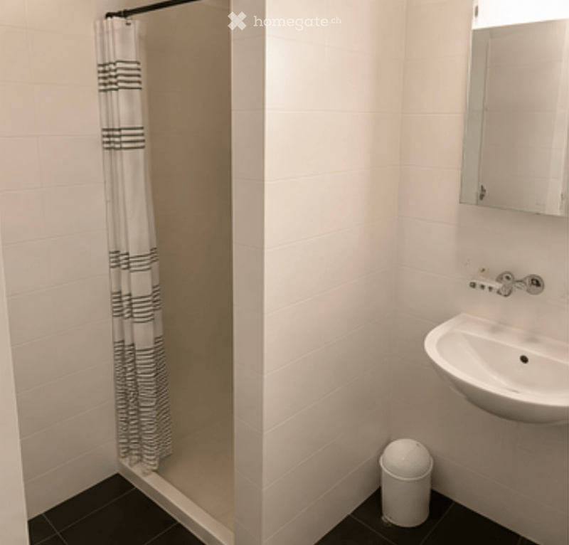 Modern bathroom with white tiled walls, black tiled floor, a corner shower with a black and white striped curtain, a white sink with a mirror above it, and a small white trash can.
