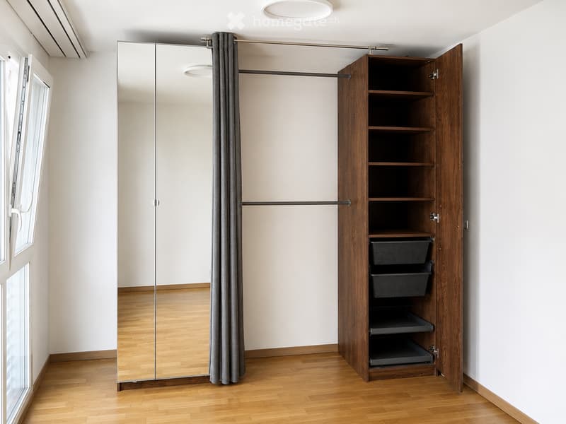 Empty room with wooden flooring, large mirrored closet with sliding doors, open wooden shelving unit with baskets, and a window with blinds.