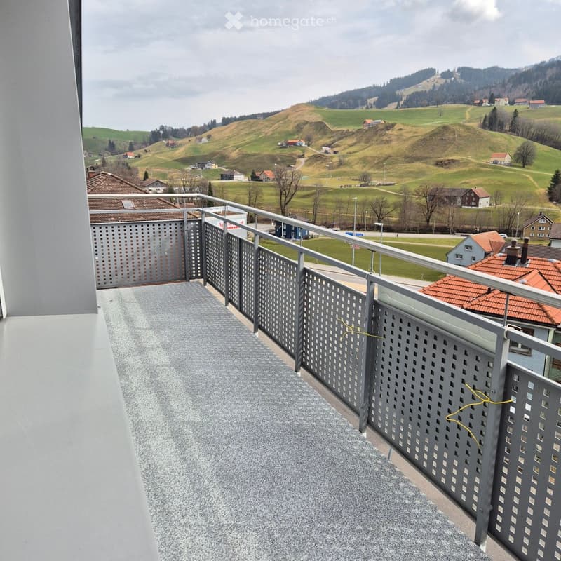Balcony with grey gravel flooring, metal railing with perforated panels, overlooking a rural landscape with rolling hills and scattered houses.