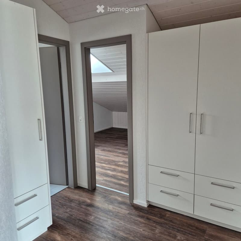 Empty room with white walls, wooden flooring, and built-in white wardrobes with silver handles. A doorway leads to another room with a skylight.