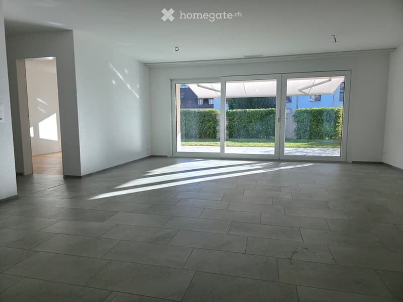 Empty room with tiled floor, white walls, large sliding glass doors leading to a garden, and natural light streaming in.