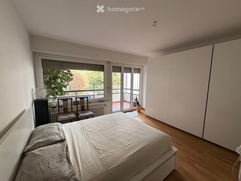 Bright bedroom with wooden flooring, large window leading to a balcony, bed with pillows, two chairs, small table, lamp, radiator, and potted plant on balcony.