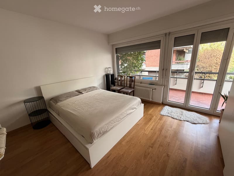 Modern bedroom with wooden flooring, large bed with white bedding, nightstand, chairs, radiator, and a view to a balcony.