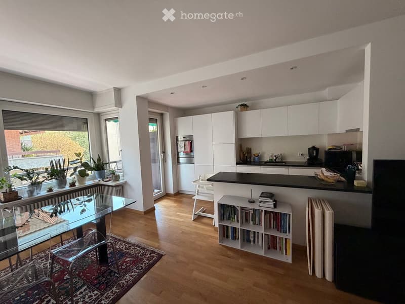 Modern living and dining area with a glass table, chairs, bookshelf, and kitchen area with cabinets, refrigerator, microwave, and sink.