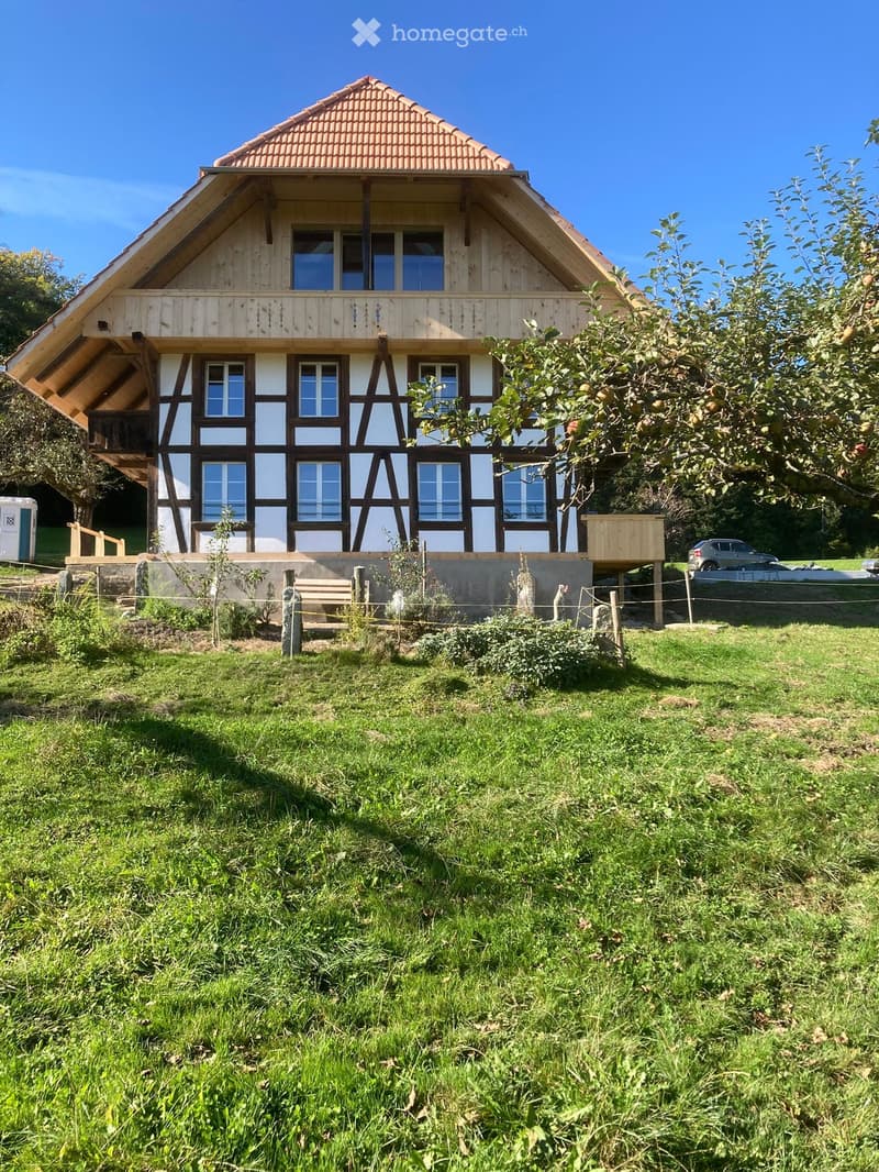 Two-story wooden house with white and brown exterior, large windows, and a raised ground floor. The house features a tiled roof and is surrounded by a grassy yard with trees and a parked car.